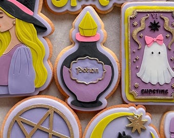 Puede incluir: Surtido de galletas con temática de Halloween. Los diseños incluyen una bruja, una botella de poción, un fantasma, un pentagrama, un gato negro y la palabra "Halloween". Las galletas presentan detalles en morado, amarillo, negro y dorado.