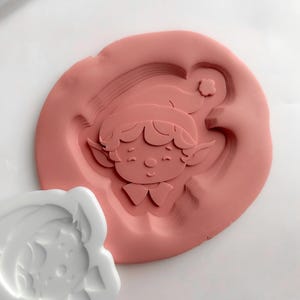May include: A pink silicone mould featuring a smiling elf face with a hat and pointed ears. A white plastic mould of the same elf face is next to the pink mould. The moulds are on a white surface.