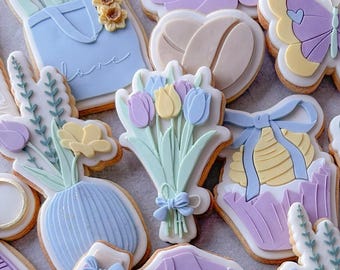 May include: A collection of decorated sugar cookies featuring spring-themed designs. The cookies are decorated with pastel colors and feature images of flowers, a coffee cup, a butterfly, a vase, and a t-shirt with the word "mum" on it.