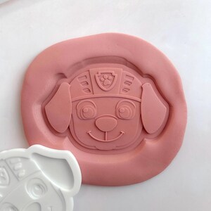 May include: A pink, circular cookie cutter depicting a cartoon dog's face, complete with large eyes, a smiling mouth, and a helmet. A white plastic cutter of the same design is next to the pink one. The design includes a paw print emblem.