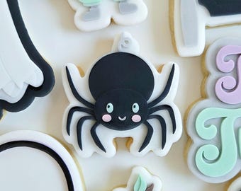 May include: A collection of Halloween-themed sugar cookies. The cookies feature various designs, including a black spider with a smiling face, a purple trick-or-treat basket, and a ghost. Other cookies depict a witch's broom and a bat.