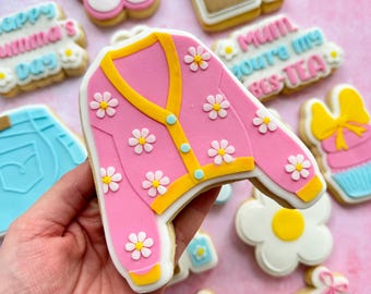 May include: A set of decorated sugar cookies with a pink cardigan cookie, a blue jean cookie, a tea bag cookie, a flower cookie, a cookie with the text "Mum you're my bes-tea", a cupcake cookie, and a banner cookie with the text "Mum".