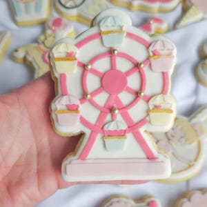 May include: A pink and white decorated sugar cookie shaped like a Ferris wheel with five pastel-coloured carriages. The Ferris wheel has a pink rim and gold beads around the wheel.