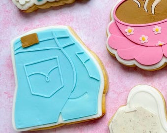 May include: A set of six decorated sugar cookies. The cookies are decorated with various designs, including a pair of blue jeans, a tea bag, a cup of tea, and a pink shirt with a yellow collar. The cookies are arranged on a pink background.