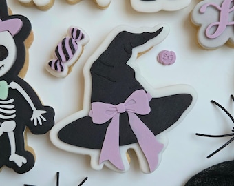 Puede incluir: Una colección de galletas con temática de Halloween. Las galletas incluyen un sombrero de bruja negro con un lazo rosa, un esqueleto, un murciélago, un zombi, un fantasma, un caramelo y una calabaza. Decoradas con glaseado negro, blanco, gris y rosa.