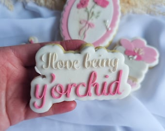 May include: A white cookie with pink icing and the text "I love being y'orchid" in white icing.