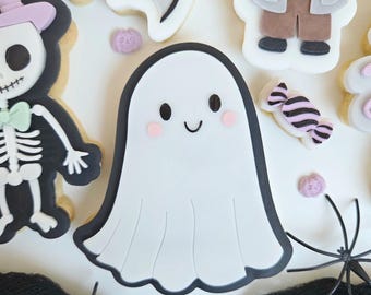 May include: A collection of Halloween-themed cookies. The cookies include a white ghost with a black outline and rosy cheeks, a skeleton, a bat, and a Frankenstein's monster. Decorative spiders and candy are also present.