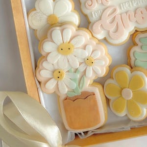 May include: A box of decorated sugar biscuits with a floral theme. The biscuits feature white and yellow daisies, a flower pot, and the text "Thank you for helping me grow". The biscuits are decorated with white, yellow, and light pink icing.