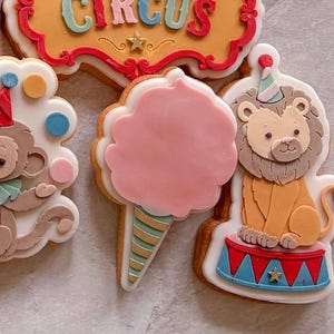 May include: A collection of decorated sugar cookies featuring a rectangular sign with the word CIRCUS in blue and red, a monkey with colourful dots, a pink cotton candy cone, and a lion wearing a party hat sitting on a red and blue circus pedestal.