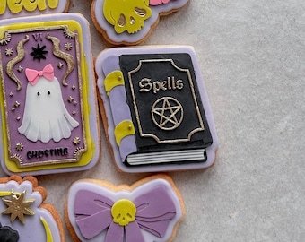 Puede incluir: Surtido de galletas con temática de Halloween con diseños intrincados. Incluye una galleta con forma de libro de hechizos, un fantasma, un lazo con una calavera y otras formas espeluznantes. Las galletas presentan detalles en morado, negro, amarillo y dorado.