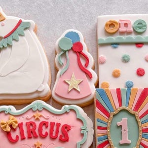 May include: A collection of five decorated sugar biscuits with a circus theme. The biscuits feature a white bear, a pink party hat with a gold star, a rectangular biscuit with the word one, a pink sign with the word CIRCUS, and a sunburst design.