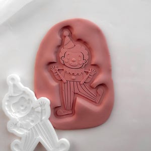 May include: A pink silicone mould featuring an embossed clown design with a hat, ruffled collar, and striped trousers, accompanied by a white plastic cutter that mirrors the same clown figure in a standing pose with arms raised.