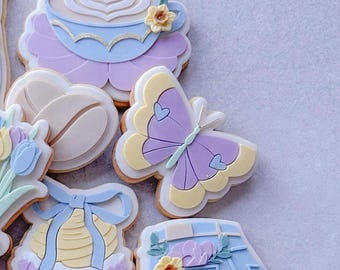 May include: Assortment of decorated sugar cookies in pastel colors. The cookies include a butterfly, coffee cup, coffee beans, cupcake with a bow, and a purse. The butterfly is purple and yellow with heart details. The coffee cup has a floral design.