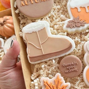 May include: A collection of decorated sugar cookies with autumn themes. The cookies feature a boot, a leaf, a pumpkin spice season cookie, and a cookie with the word "Autumn" on it.