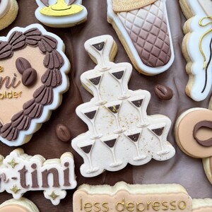 May include: A collection of decorated sugar cookies with coffee and martini themes. The cookies feature brown, white and gold icing with various designs, including a coffee cup, a martini glass, and the phrase "less depresso more espresso".