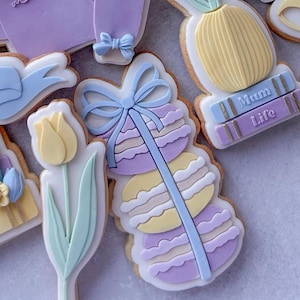May include: A set of decorated sugar cookies with pastel colours. The cookies feature a flower, a stack of macarons with a blue ribbon, a sweater with the word "mum" on it, and a vase with flowers on top of a book that says "Mum Life".