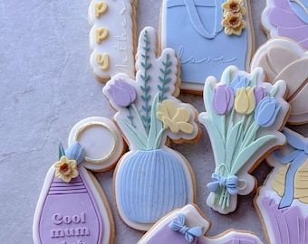 May include: Assortment of decorated sugar cookies with various designs, including flowers, a vase, a shopping bag with the word "love", a key, a purple cookie with the text "Cool mum club", and a purple shirt with the word "mum".