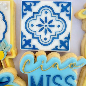 May include: Close-up of decorated sugar cookies. One cookie features a blue and white tile design. Another cookie has the words "Ciao MISS" in blue and gold. Other cookies have lemon and floral designs.