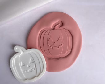 May include: A white plastic pumpkin-shaped mold with a carved jack-o'-lantern face. The mold is sitting on a pink silicone baking mat with a matching pumpkin design.