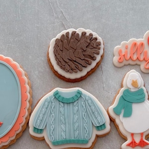 May include: Selection of decorated biscuits. One biscuit is shaped like a pine cone, another like a light blue sweater, and another like a duck wearing a teal scarf. The word "silly" is written on one biscuit in orange icing.