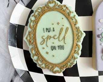 Puede incluir: Una galleta decorada en verde y dorado con el texto "I put a spell on you" en un marco dorado.