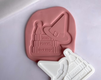 Puede incluir: Un molde de silicona blanco para hacer manualidades de resina. El molde presenta un sombrero de bruja, una botella y una pila de libros con las palabras "Spells, Potions, Black Magic".