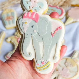 May include: A decorated sugar cookie shaped like an elephant wearing a pink and white hat, holding a ball with gold and pink accents. The cookie is decorated with pink, white, and gold icing.