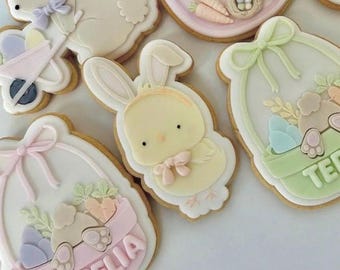 May include: A set of five Easter-themed sugar cookies decorated with pastel icing. The cookies feature a bunny pushing a wheelbarrow, a chick with a bow, a basket with pastel eggs, and two baskets with the names "Aurelia" and "Teddy".