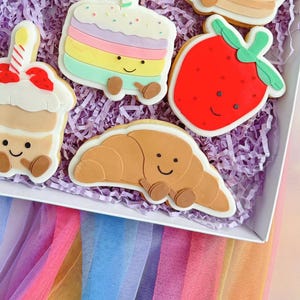 May include: A selection of decorated biscuits, including a rainbow cake, a strawberry, a croissant, and a cupcake, all with smiling faces. The biscuits are on purple paper, inside a white box, with a colourful fabric background.