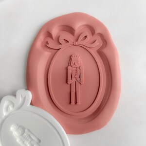 May include: Two crafting molds, one white and one dusty rose. Each features a detailed design of a nutcracker soldier within an oval frame, topped with a bow. These molds are likely for creating decorative items, perfect for festive crafts.
