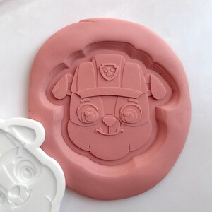 May include: A pink silicone mould featuring a cartoon dog character with a hat and badge. Beside it is a white, dog-shaped cookie cutter. The mould has a circular shape with the dog's face in the centre, ideal for baking or crafting.