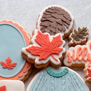 May include: A selection of decorated biscuits with autumn-themed designs. The biscuits feature a maple leaf, a pinecone, a sweater, and a plate with a leaf. The biscuits are decorated with orange, brown, and blue icing.
