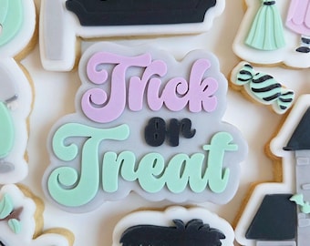 May include: A collection of Halloween-themed sugar cookies. The cookies feature designs like a sign that says "BOO", "Trick or Treat" lettering, a Frankenstein's monster, a witch, a ghost, and a bat. The cookies are decorated with pastel colors.