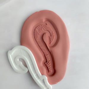 May include: A white plastic mould and a pink clay mould, both featuring a curved hook design with detailed line patterns. The hook shape is reminiscent of a traditional fish hook or a stylized symbol.