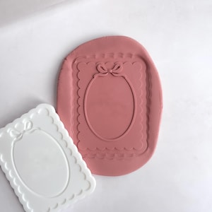 May include: Two silicone moulds for making polymer clay jewellery. The mould on the left is white and the mould on the right is pink. Both moulds feature a rectangular frame with a scalloped edge and a bow at the top. The frame contains an oval cutout.