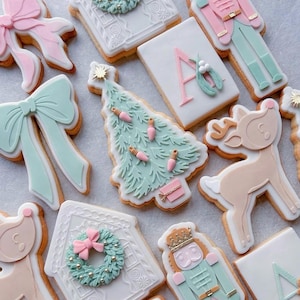 May include: A collection of Christmas-themed sugar cookies decorated with various designs, including a Christmas tree, a reindeer, a nutcracker, a gingerbread house, and a bow. The cookies are all white with pastel green, pink, and gold accents.