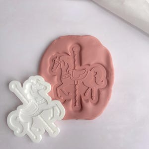 May include: A white plastic carousel horse cookie cutter with a pink silicone mould showing the impression of the horse.