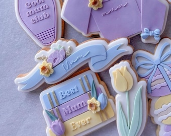 May include: A set of decorated sugar cookies with various designs, including a blue and purple cookie with the text "Cool Mum Club", a purple cookie shaped like a sweater with the text "Mum", a blue and yellow cookie with the text "Best Mum Ever", a yellow tulip cookie, and a purple cookie with a bow.