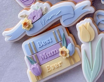 May include: A set of decorated sugar cookies with a blue ribbon banner that says "Mum", a stack of books that says "Best Mum Ever", and a yellow tulip.