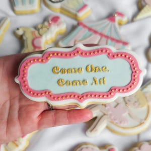 May include: A light blue and pink decorated cookie with gold lettering that reads "Come One, Come All".