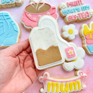 May include: A set of decorated sugar cookies with various designs, including a tea bag, a cup of tea, a daisy, a banner that says "mum", and a cookie that says "Mum, you're my bes-tea".