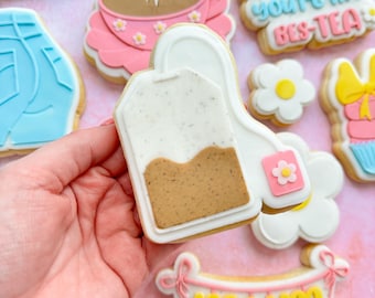 May include: A set of decorated sugar cookies with various designs, including a tea bag, a cup of tea, a daisy, a banner that says "mum", and a cookie that says "Mum, you're my bes-tea".