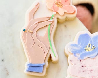 May include: A hand-painted sugar cookie decorated with a pink flower and a green stem. The cookie is shaped like a hand holding the flower. The hand is painted in a light pink color and has a blue bracelet on the wrist.