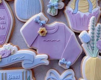 May include: A set of decorated sugar cookies with various designs, including a vase of flowers, a cupcake, a sweater with the word "mum" on it, and a stack of books with the words "Best Mum Ever" and "Mum Life".