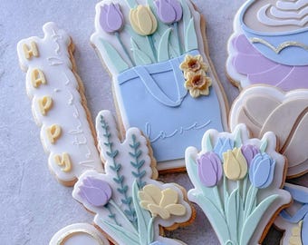May include: A collection of decorated sugar cookies featuring floral designs, a coffee cup, and the word "Happy" in yellow letters. The cookies are decorated with pastel colors, including blue, yellow, and purple.