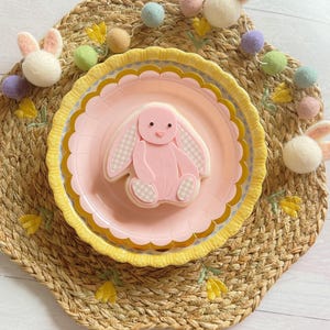 May include: An Easter table setting with a pink bunny cookie on a pink plate with a gold rim, on a yellow scalloped plate. The plates are on a woven placemat, with felt bunny decorations and a garland of pastel felt balls. The setting evokes a spring theme.