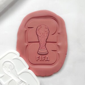 May include: A white plastic cookie cutter and a pink pressed dough shape featuring the number 26 with the FIFA World Cup trophy design embossed in the centre. The trophy is depicted with a globe at the top and the text FIFA printed below it.