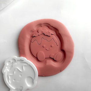 May include: A white plastic cookie cutter shaped like a Christmas pudding with a smiling face and holly leaves, positioned next to a matching terracotta-coloured clay impression that shows the embossed details of the design clearly.
