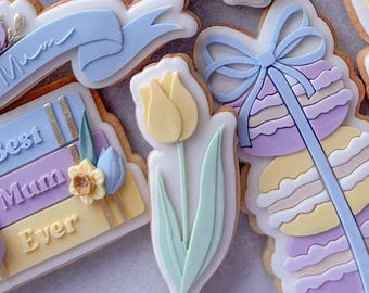 May include: A set of decorated sugar cookies with various designs, including a stack of books with the text "Best Mum Ever", a blue ribbon with the word "Mum", a yellow tulip, and a stack of macarons with a blue ribbon.