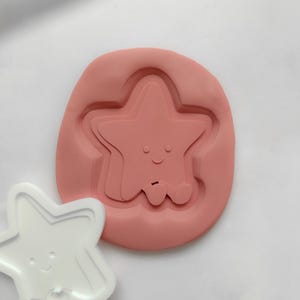 May include: A pink silicone mould and a white plastic star-shaped cookie cutter. The mould features a smiling star design with arms and legs. The cookie cutter has a similar design, with a smiling star.